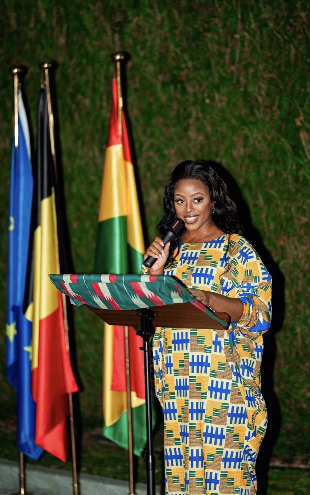 Queenie Kessie speaking at a lectern in a kente-print dress, with the flags of Ghana, Belgium and the European Union behind her